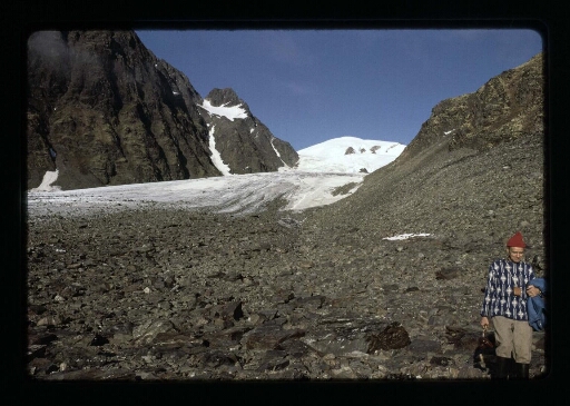 Un homme debout  portant un bonnet rouge; derrière lui le glacier de la Baie du Roi - mission CNRS 1965