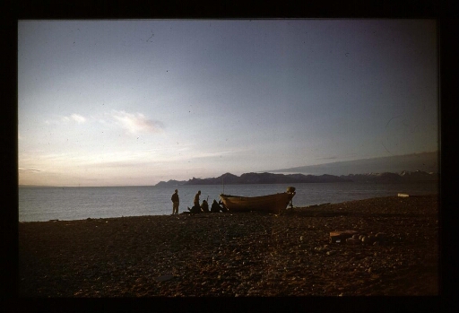 Des hommes sur la rive d'un fjord près d'une barque en bois sous un couché de soleil - mission CNRS 1966