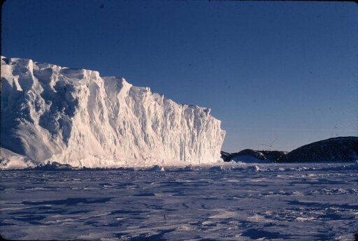 Un gros iceberg tabulaire immobilisé par la banquise. En arrière-plan on aperçoit les installations de la base Dumont d'Urville.