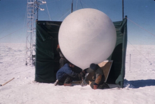 Sondage météo à la station Charcot : trois hommes préparent le ballon derrière un pare-vent, au pied de la tour météo.