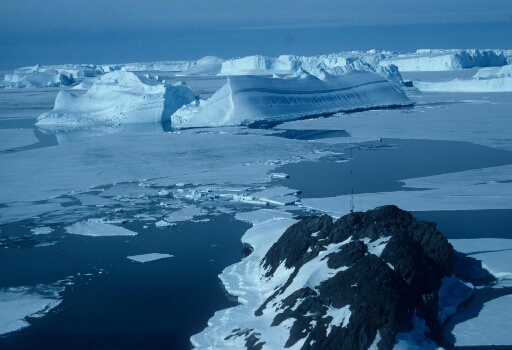 Survol du rocher du Débarquement surmonté par le mât du programme IAGO. Banquise en partie disloquée, icebergs tabulaires et deux icebergs retournés.
