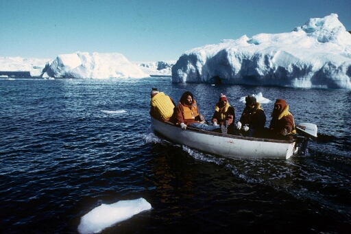 Cinq hommes à bord d'un canot naviguant en mer libre au milieu de petits icebergs. En arrière plan, le glacier de l'Astrolabe.