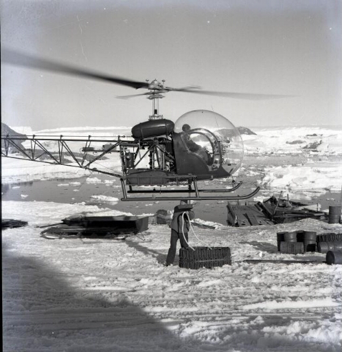 Transport des matériels en "Sling" : un homme accroche une chenille de véhicule sous l'hélicoptère Bell 47 G2.