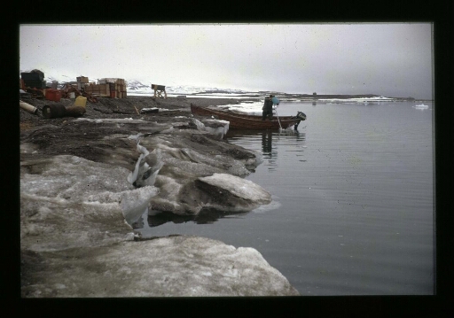 Un homme debout près d'une barque et de matériels de l'équipe sur une rive du fjord - mission CNRS 1966