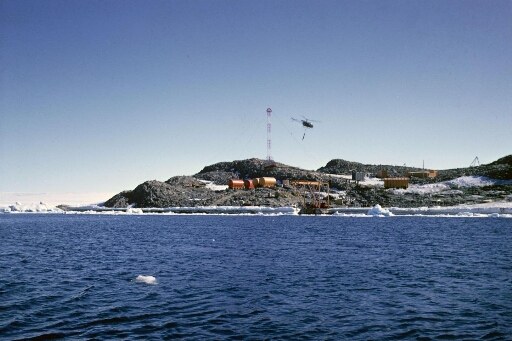 Le sud de l'île des Pétrels vu de la mer. L'hélicoptère Alouette 2 en opération de transport. En arrière-plan, le pylône du sondeur ionosphérique.