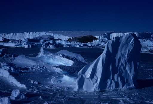 Hummocks ancrés dns la banquise non loin d'un îlot et, en arrière-plan, un grand iceberg tabulaire.