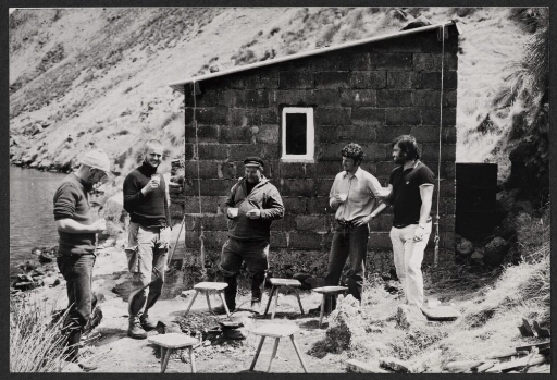 Cinq hommes, un verre à la main, autour d'un barbecue devant une cabane