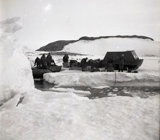 La barge accostée à la glace de mer. Les hommes chargent les caisses débarquées sur un traîneau tiré par un weasel.