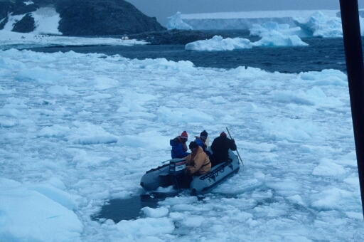 Quatre hommes à bord d'un zodiac naviguant dans un pack très dense de l'archipel. A l'arrière-plan, un grand iceberg tabulaire.