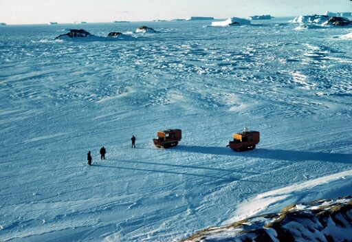 Sur la glace de mer, deux weasels, trois hommes à pied. Mer gelée à perte de vue, de nombreux icebergs et quelques ilots de l'archipel.