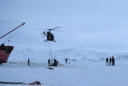 Déchargement du Thala Dan sur la banquise à l'aide de l'hélicoptère Alouette 2. Une dizaine d'opérateurs. Les icebergs proche du continent.