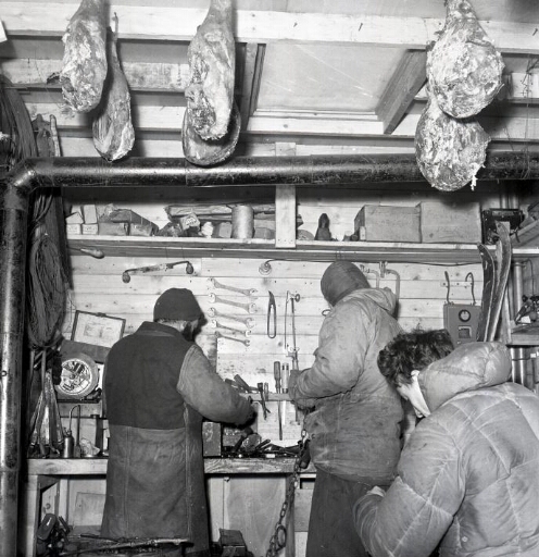 Trois hommes travaillent dans l'atelier. Les jambons secs sont pendus à une poutre.