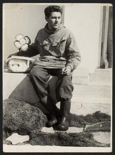 Un homme assis sur les marches d'une habitation, fumant une cigarette. Une pierre et une fleur à sa droite