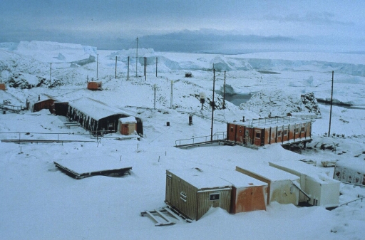 Hiver 1988, l'ancienne base et la station radio sous la neige. L'antenne Caroline. Au loin, le glacier de l'Astrolabe. Ciel bas.