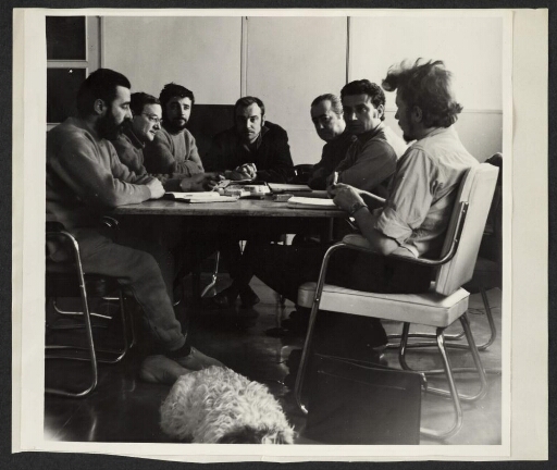 Huit hommes attablés en réunion. Un chien couché au centre.