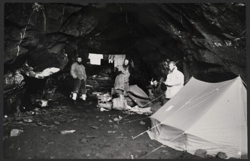 Quatre hommes dans une grotte. Une tente installée, du linge qui tend à un fil et du matériel