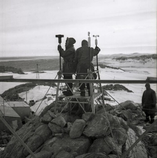 Deux hommes contrôlent les appareils météo installés sur la plateforme installée sur un relief surplombant la base.