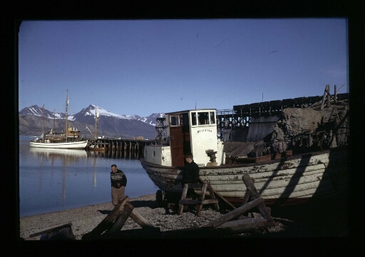 Deux hommes près d'un bateau hors de l'eau qui se trouve à coté d'un quai du port de Ny-Ålesund - mission CNRS 1963