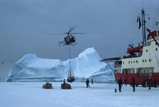 Déchargement du Thala Dan sur la banquise à l'aide de l'hélicoptère Alouette 2. Une dizaine d'opérateurs. Ciel plombé, bel iceberg.