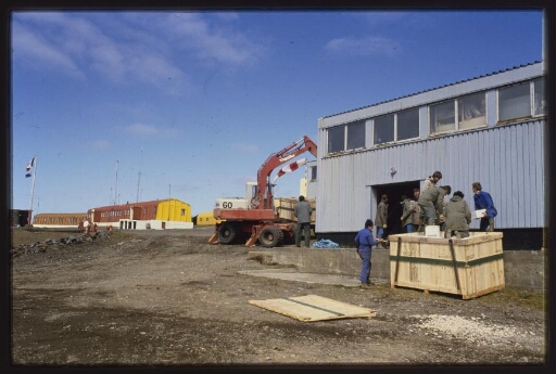 Bâtiments de la base, grue, hangar. Des hommes autour d'une caisse de transport