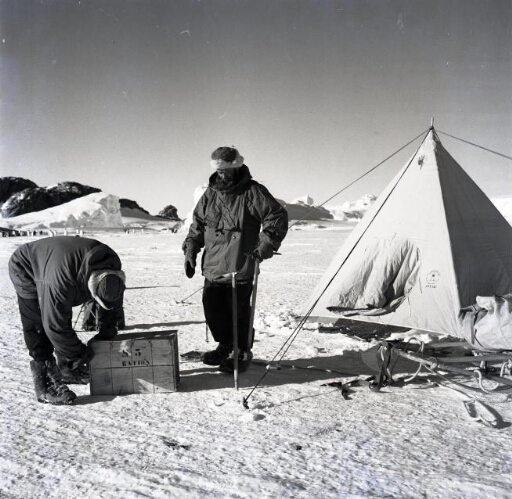 Deux hivernants devant une tente installée sur la banquise. A l'arrière-plan, des manchots empereurs.