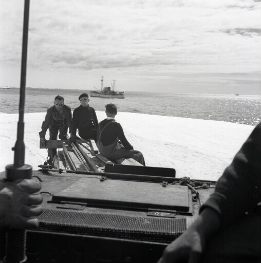 Trois hommes assis sur un convoi de matériel tracté par un weasel. Au large le Commandant Charcot au mouillage.