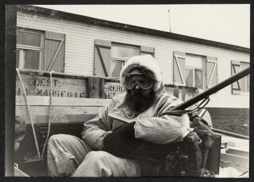 Portrait d'un homme portant une capuche, un fusil et des lunettes de neige. Arrière-plan : bâtiment