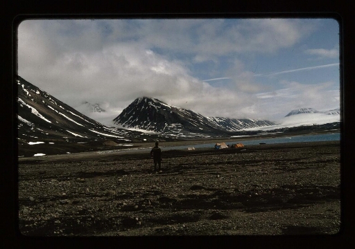 Un homme debout, derrière lui un campement sur la plage près d'un fjord dans le paysage du Spitzberg - mission CNRS 1966 - vue 1