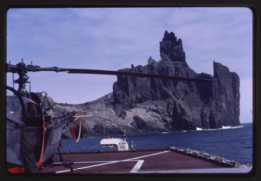 Hélicoptère orange posé sur la piste d'atterrissage d'un navire. Ile en arrière-plan. Diapo 1