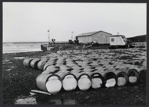 Bâtiment de type hangar au bord de la côte. Grue et barils en bois alignés au premier plan