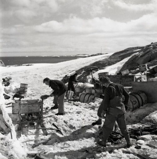 Quatre hommes s'emploient au rangement du matériel débarqué. Au loin, la mer libre, beau temps calme.