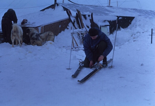Le bâtiment principal de Port-Martin, en partie enseveli sous la neige. Un homme chausse ses skis, un autre s'occupe des chiens.