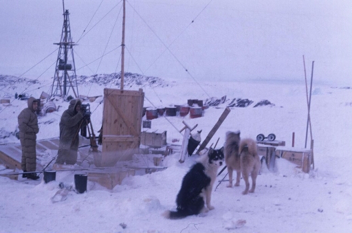 Travaux extérieurs à Port-Martin : deux hommes dont l'un, au théodolite, suit le ballon-sonde météo. Deux chiens, temps hivernal.