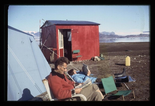 Jean Corbel assis sur une chaise de camping dans la base Corbel à coté d'une femme qui lit, allongée sur un transat - mission CNRS 1964