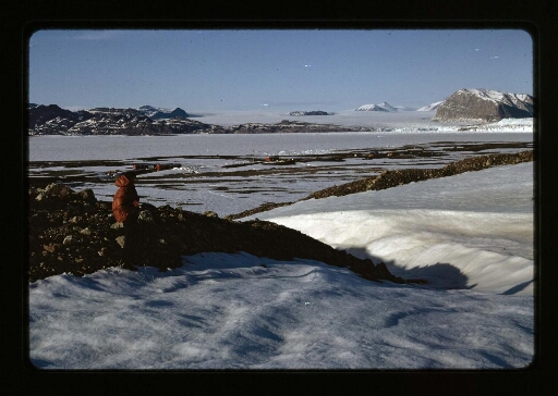 Un homme devant un décor enneigé, au loin des bâtiment,un glacier et un fjord gelé - mission CNRS 1966