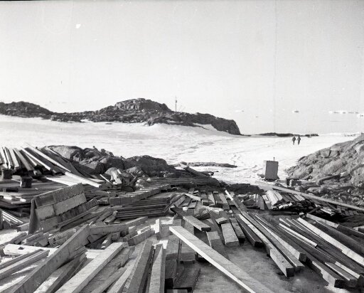 Les bois de construction de la base. Au loin deux hommes et, derrière le rocher, la mâture du Commandant Charcot.
