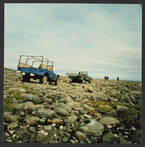 Deux véhicules de type jeep au premier-plan avec un chauffeur. Trois hommes marchant au loin. Paysage de rochers
