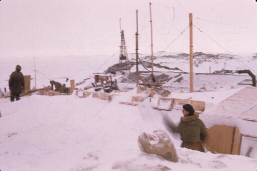 Port-Martin sous la neige dont on ne voit que le toit et le pylône de l'éolienne. Trois hommes s'affairent sur les caisses de matériels.