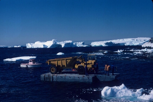 Transport d'un "dumper" sur un ponton pour le chantier de la piste du Lion. Mer libre. Deux beaux icebergs en l'arrière-plan.
