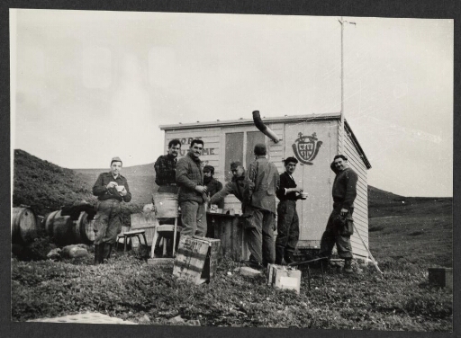 Un groupe d'hommes mangent un repas en plein air devant la cabane de Port Douzième au pied du pouce sur la presqu’île Ronarc'h