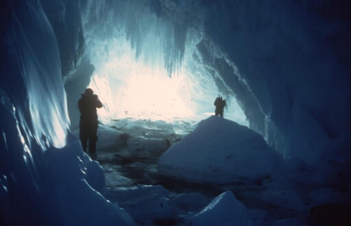 En fin d'hivernage, la banquise encore solide permet à ces deux hivernants d'explorer une grotte dans un iceberg. Lumière bleue caractéristique et stalactites.