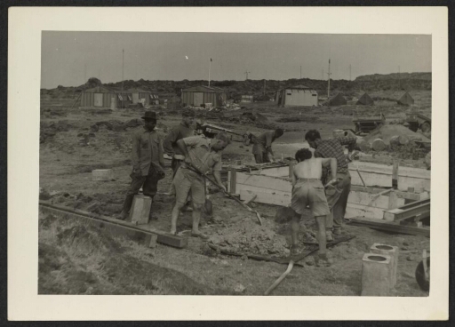 Des hommes sur un chantier. À l'arrière plan des bâtiments hangars et des tentes.