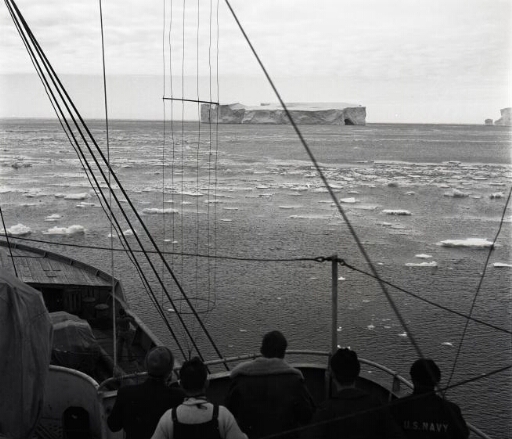 Depuis la passerelle, cinq hommes contemplent un iceberg tabulaire. Mer calme chargée de floes.