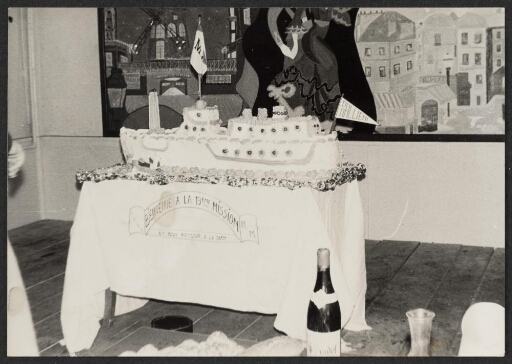 Banquet avec un gâteau de bienvenue en forme de navire. Inscription sur la nappe : "M.M. Bienvenue à la 19ème mission et bon retour à la 18ème".