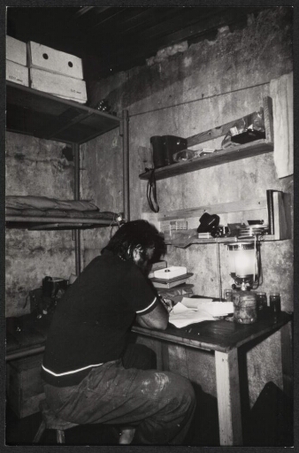 Un homme assis à une table, dans un bâtiment, écrivant, des couchettes en arrière-plan