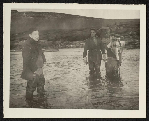 Trois hommes munis de bottes ont les pieds dans l'eau. Un des hommes tient un appareil de forage