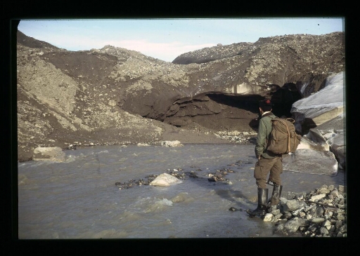 Un homme debout de dos, les pieds dans l'eau qui regarde des éboulis de roche sur un plan d'eau - mission CNRS 1966