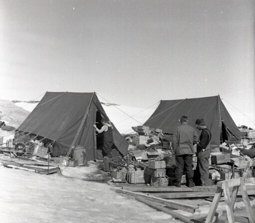Le chantier de la base. Deux tentes sont dressées au milieu d' un amoncellement de caisses. Trois hommes.