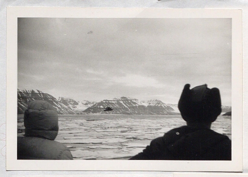 Deux hommes de dos observe un hélicoptère qui survole la mer ou flottent des morceaux de glace