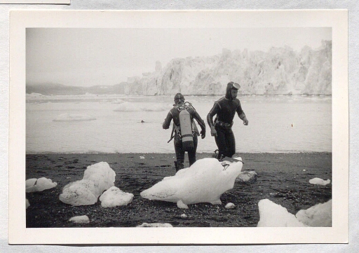 Deux hommes en tenue de plongée sur le bord de l'eau en face d'un glacier